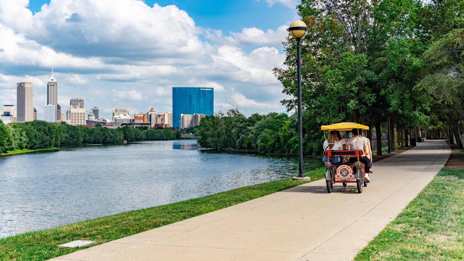 people riding a vehicle beside a river, skyline at the back and trees on the side